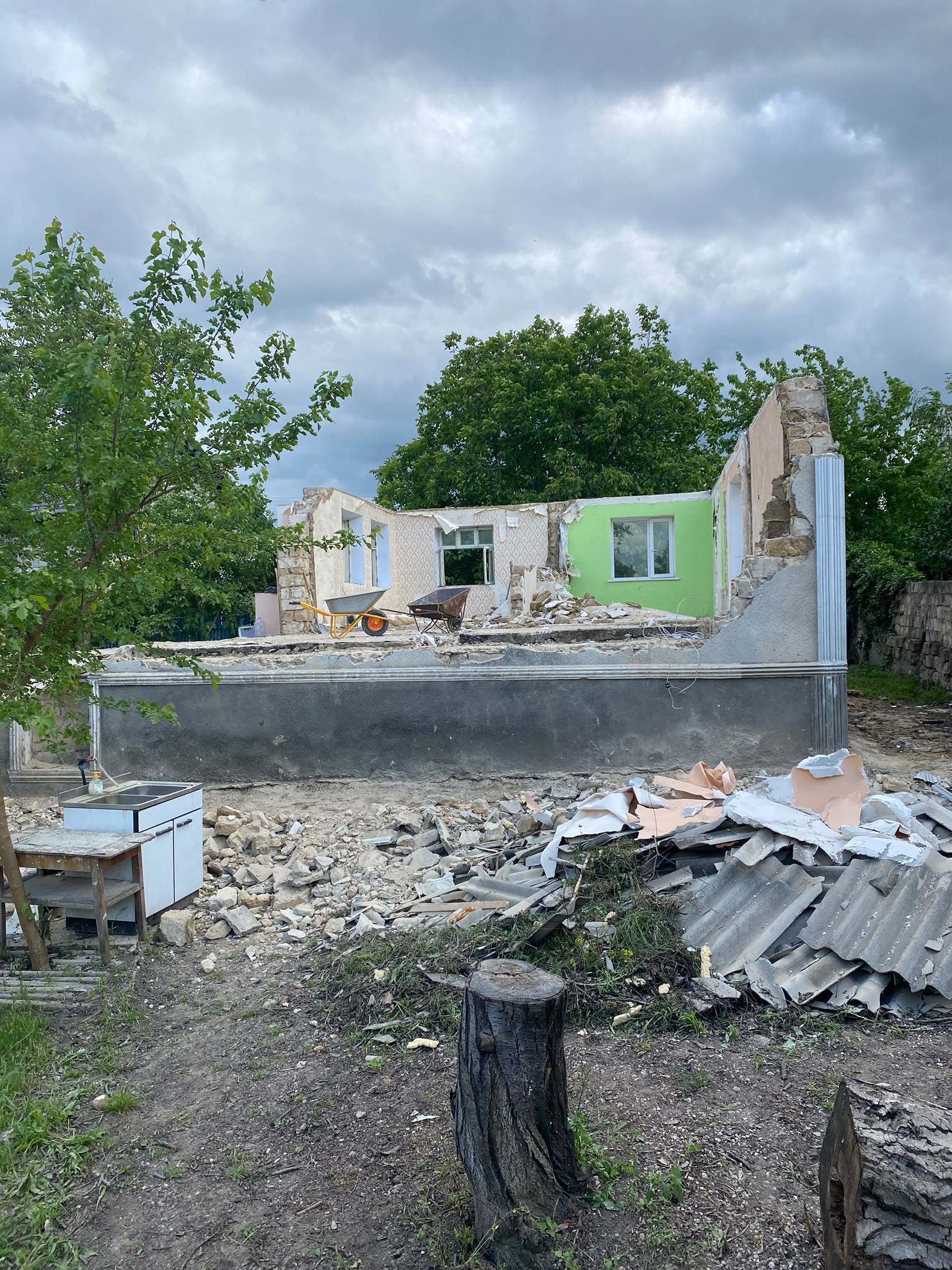 Casă parțial demolată, cu moloz, foi ondulate de acoperiș și un buștean tăiat în curte